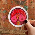 Someone holding the Libra Zodiac 3 Inch Sticker in front of a wooden background.