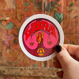 Someone holding the Libra Zodiac 3 Inch Sticker in front of a wooden background.