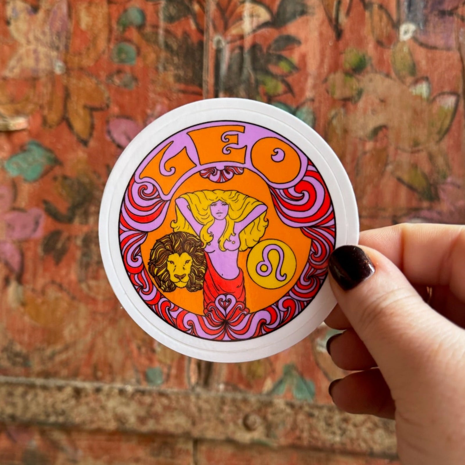 Someone holding the Leo Zodiac 3 Inch Sticker in front of a wooden background.