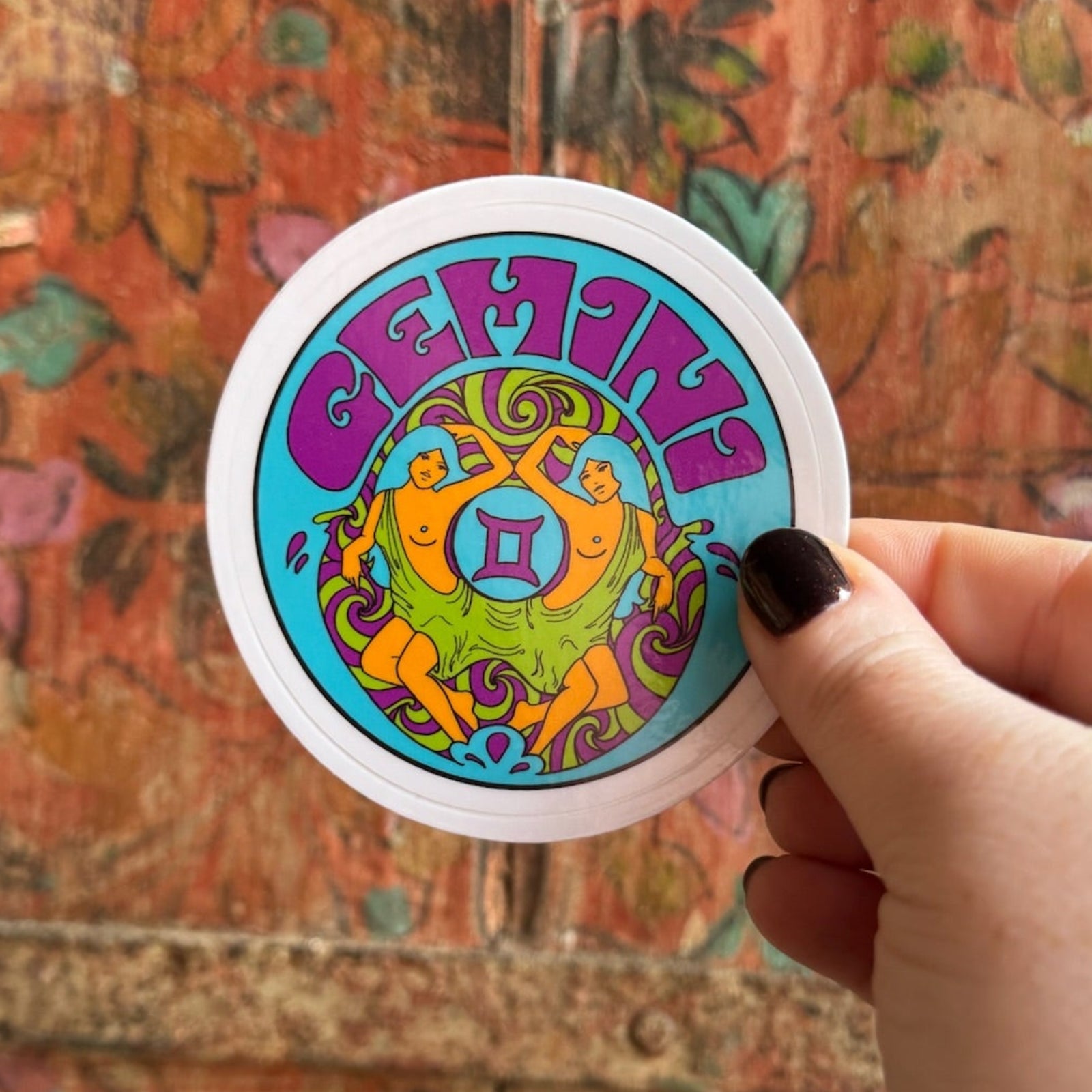 Someone holding the Gemini Zodiac 3 Inch Sticker in front of a wooden background.