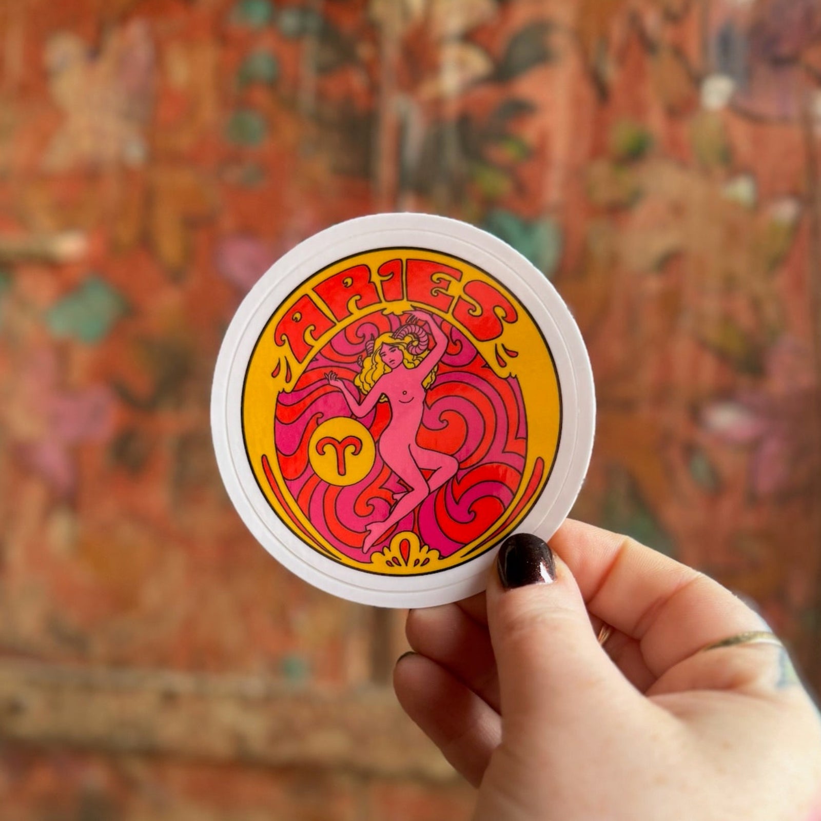 Someone holding the Aries Zodiac 3 Inch Sticker in front of a wooden background.
