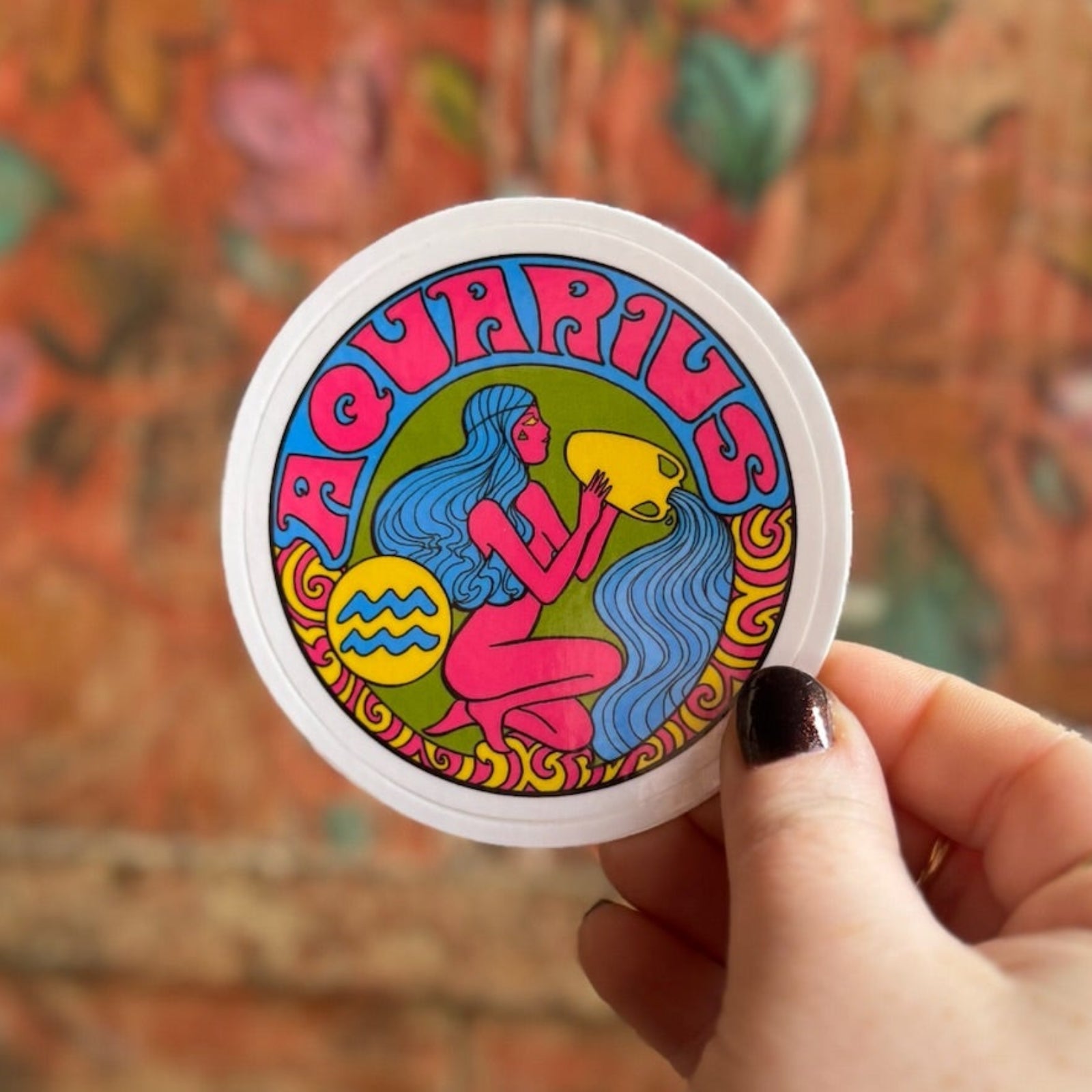 Someone holding the Aquarius Zodiac 3 Inch Sticker in front of a wooden background.