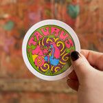 Someone holding the Taurus Zodiac 3 Inch Sticker in front of a wooden background.