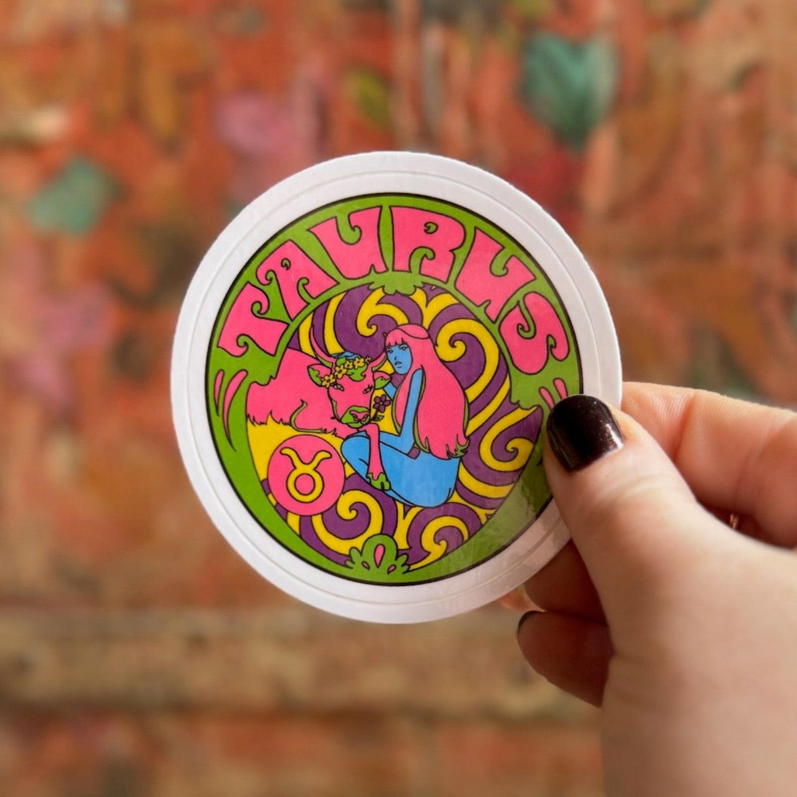 Someone holding the Taurus Zodiac 3 Inch Sticker in front of a wooden background.