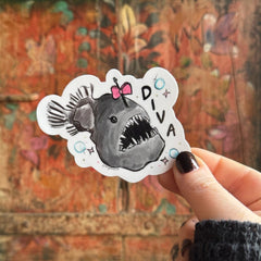 Someone holding the Anglerfish Diva Vinyl Sticker in front of a wooden background.