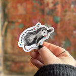 Someone holding the Socially Drained Possum Vinyl Sticker in front of a wooden background.