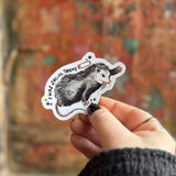 Someone holding the Socially Drained Possum Vinyl Sticker in front of a wooden background.