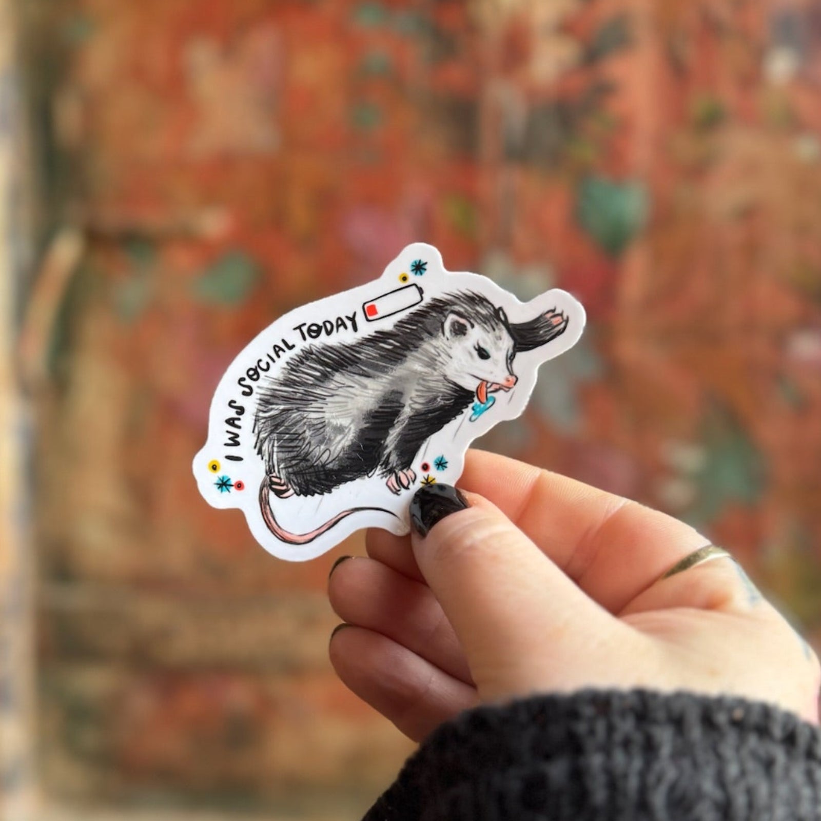 Someone holding the Socially Drained Possum Vinyl Sticker in front of a wooden background.