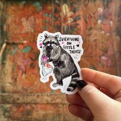Someone holding the Little Treat Raccoon Vinyl Sticker in front of a wooden background.