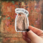 Someone holding the Pesto the Penguin Vinyl Sticker in front of a wooden background.