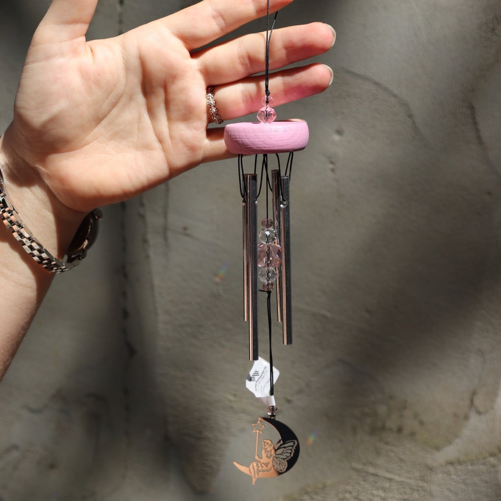 Someone holding the Fairy Fantasy Woodstock Wind Chime in front of a white cement wall.