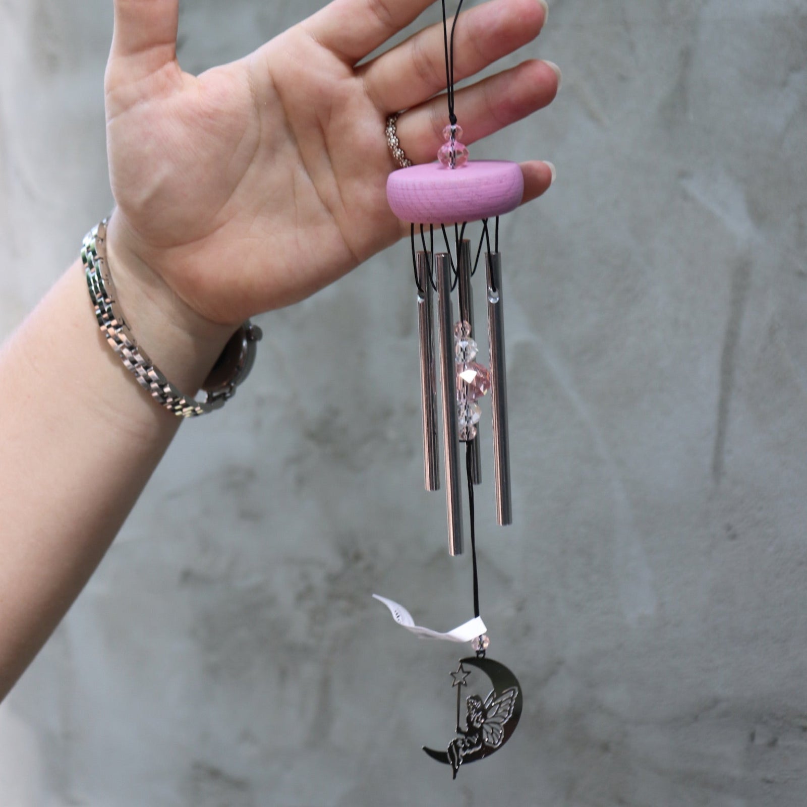 Someone holding the Fairy Fantasy Woodstock Wind Chime in front of a white cement wall.