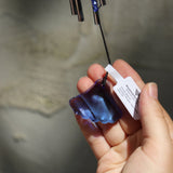 An up-close image of someone holding the Sapphire Gem Drop Wind Chime in front of a white cement wall.