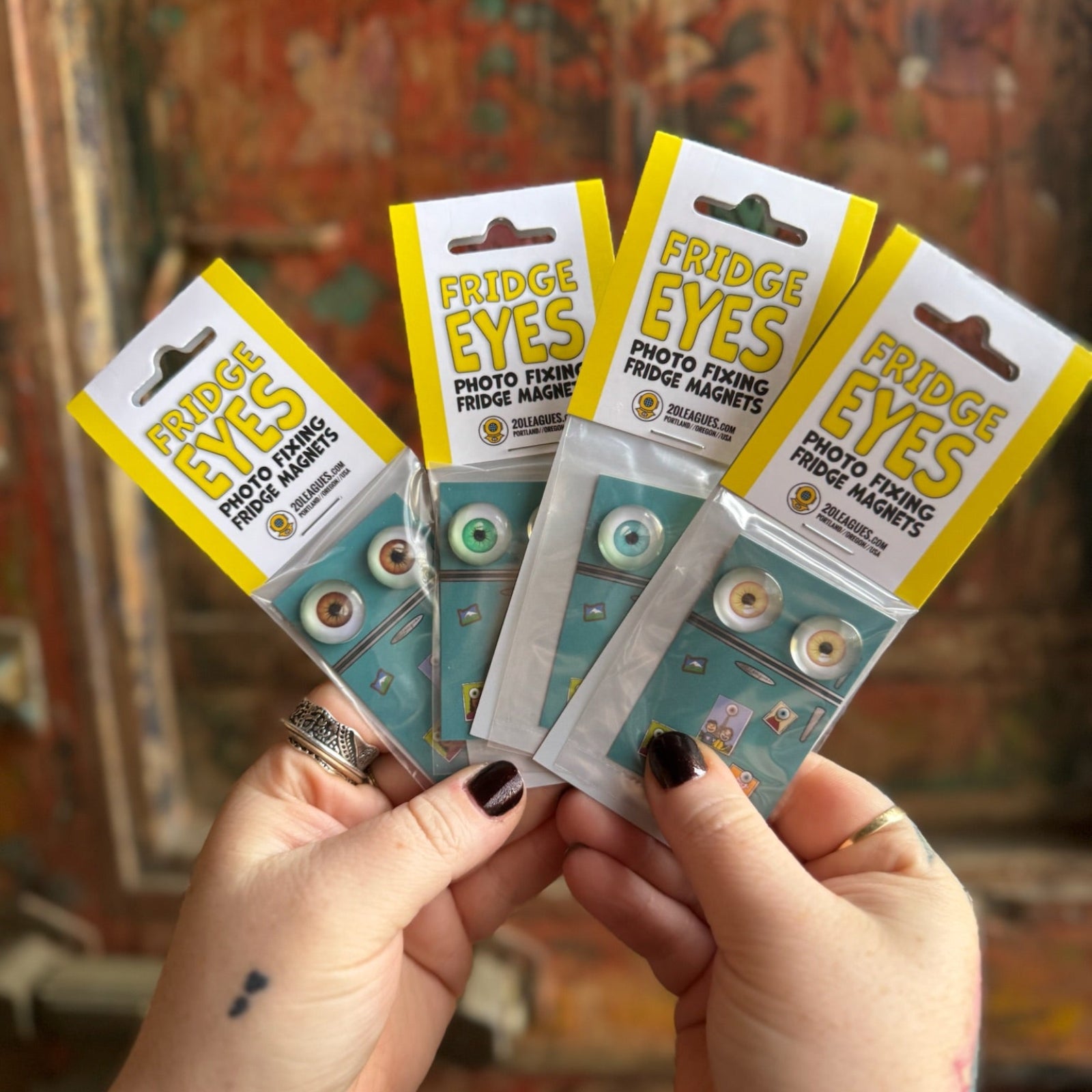 Someone holding four of the Fridge Eyes Magnets in front of a wooden background.