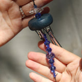 An up-close image of someone holding the Sapphire Gem Drop Wind Chime in front of a white cement wall.