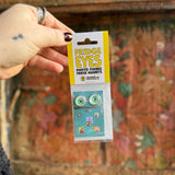 Someone holding the Fridge Eyes Magnets in front of a wooden background.