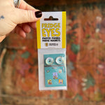 Someone holding the Fridge Eyes Magnets in front of a wooden background.