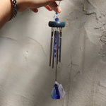 Someone holding the Sapphire Gem Drop Wind Chime in front of a white cement wall.