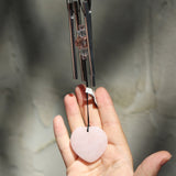 Someone holding the Rose Quartz Heart Wind Chime in front of a white cement wall.