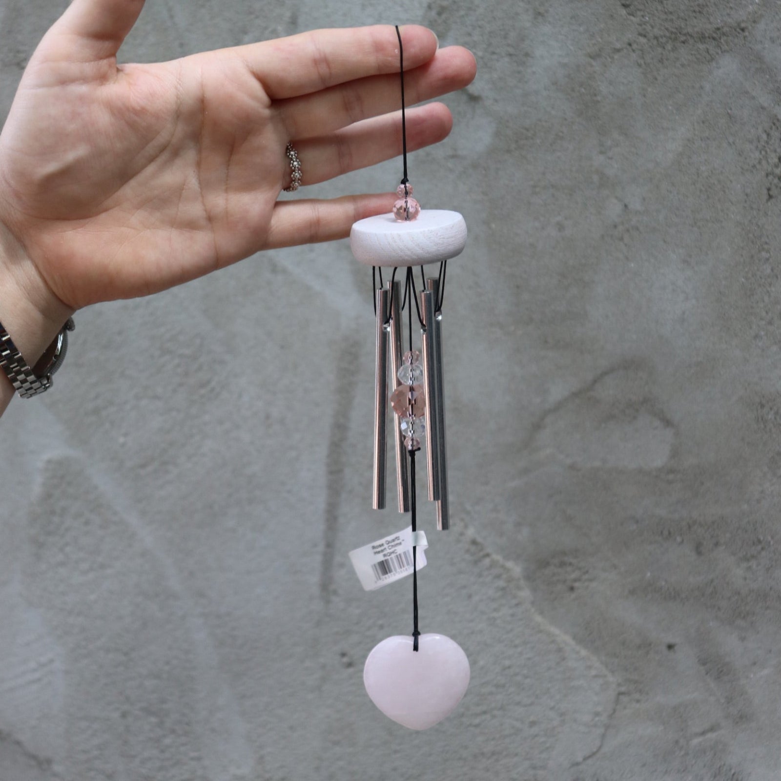 Someone holding the Rose Quartz Heart Wind Chime in front of a white cement wall.