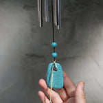 An up-close image of someone holding the accent piece on the Turquoise Woodstock Chakra Wind Chime in front of a white wall.