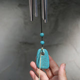 An up-close image of someone holding the accent piece on the Turquoise Woodstock Chakra Wind Chime in front of a white wall.