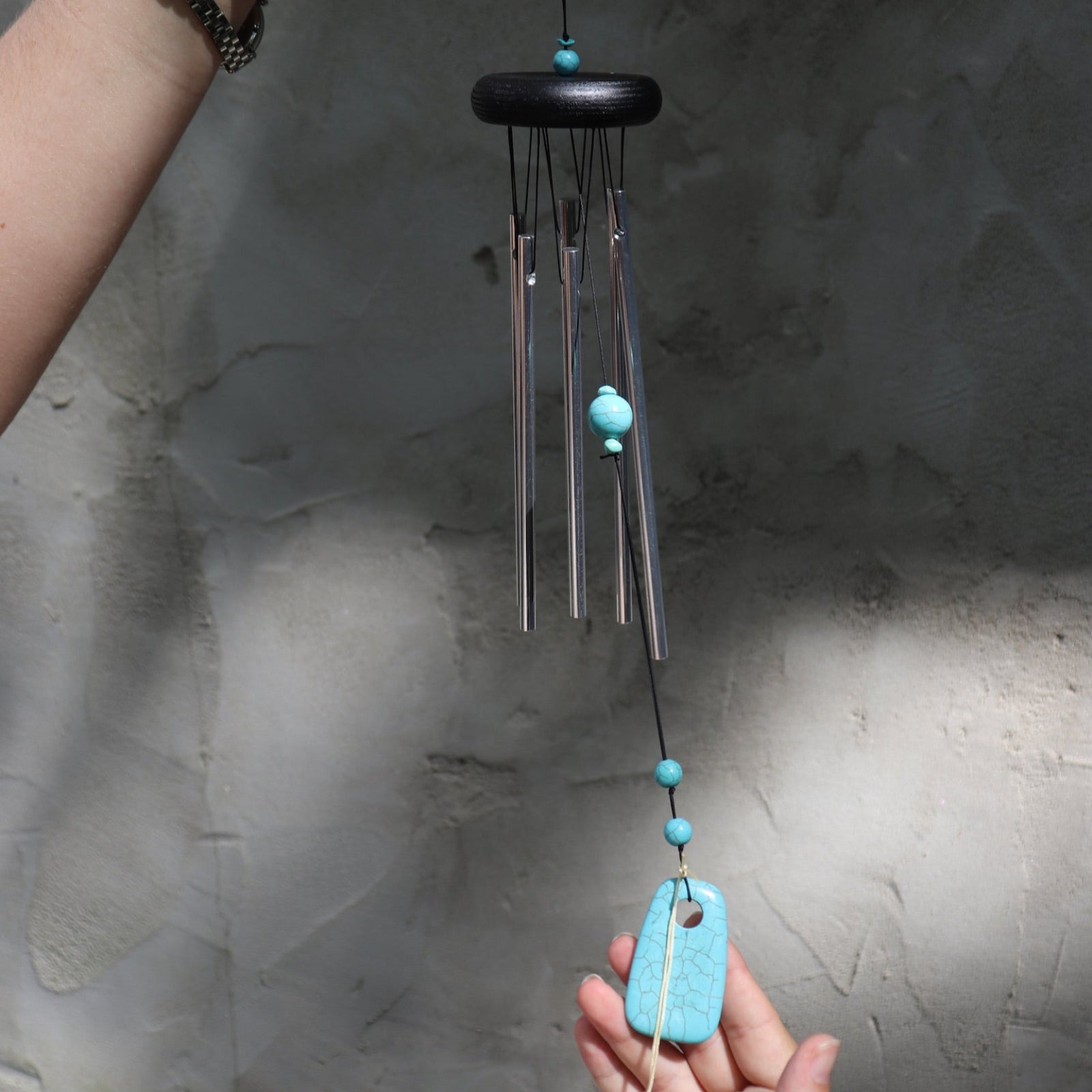 Someone holding the Turquoise Woodstock Chakra Wind Chime in front of a white wall.