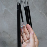 An up-close image of someone holding the Black Crystal Meditation Woodstock Wind Chime in front of a white wall.