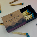 The Palo Santo Matchstick box on a pale pink and yellow background with matchsticks surrounding it.