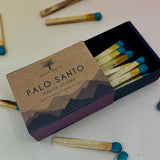 The Palo Santo Matchstick box on a pale pink and yellow background with matchsticks surrounding it.