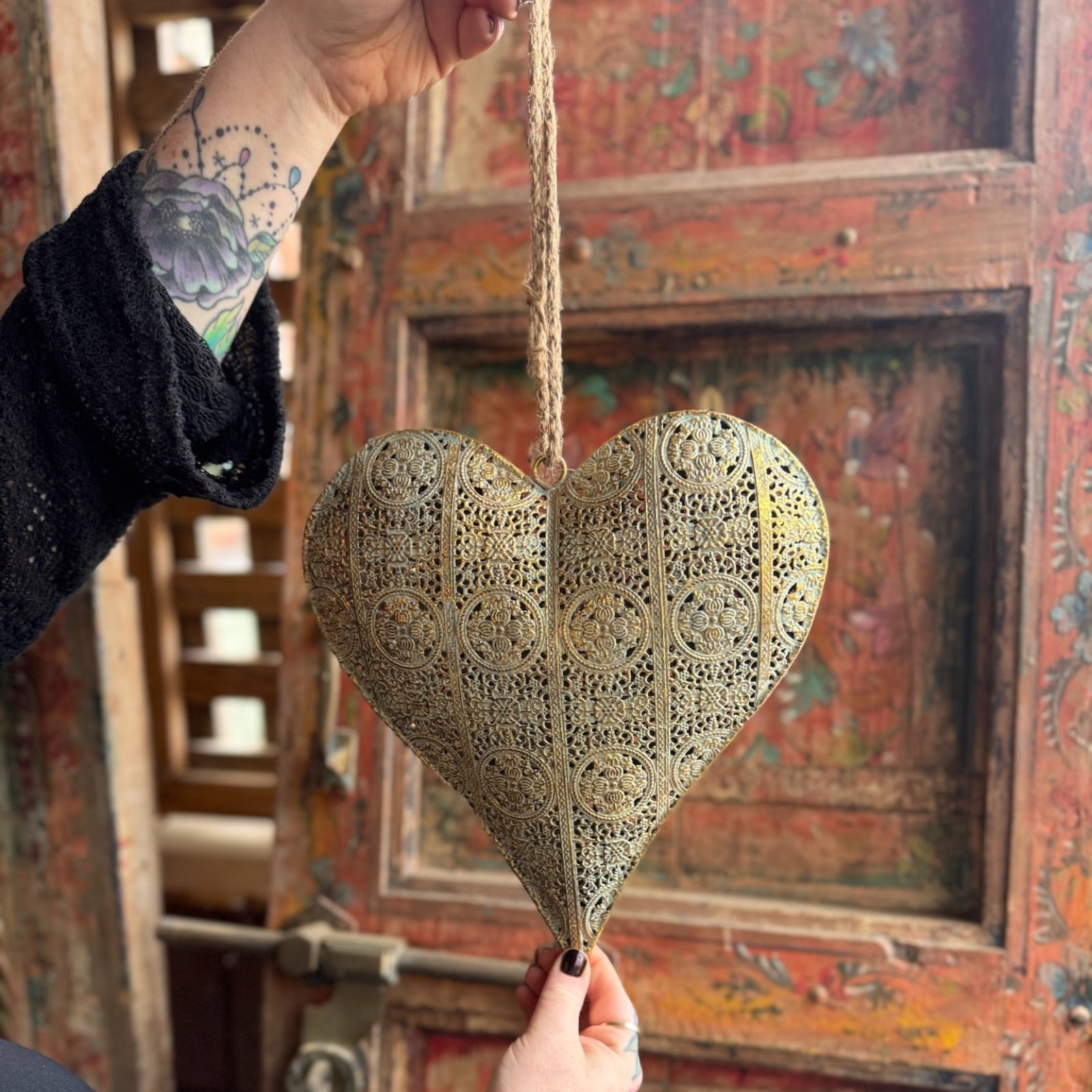 Someone holding the Ashia Gold Metal Heart Tea Light Holder in front of a wooden background, showing off the back.