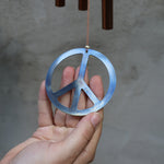 An up-close image of someone holding the metal peace sign from the Peace Woodstock Wind Chime in front of a white wall.