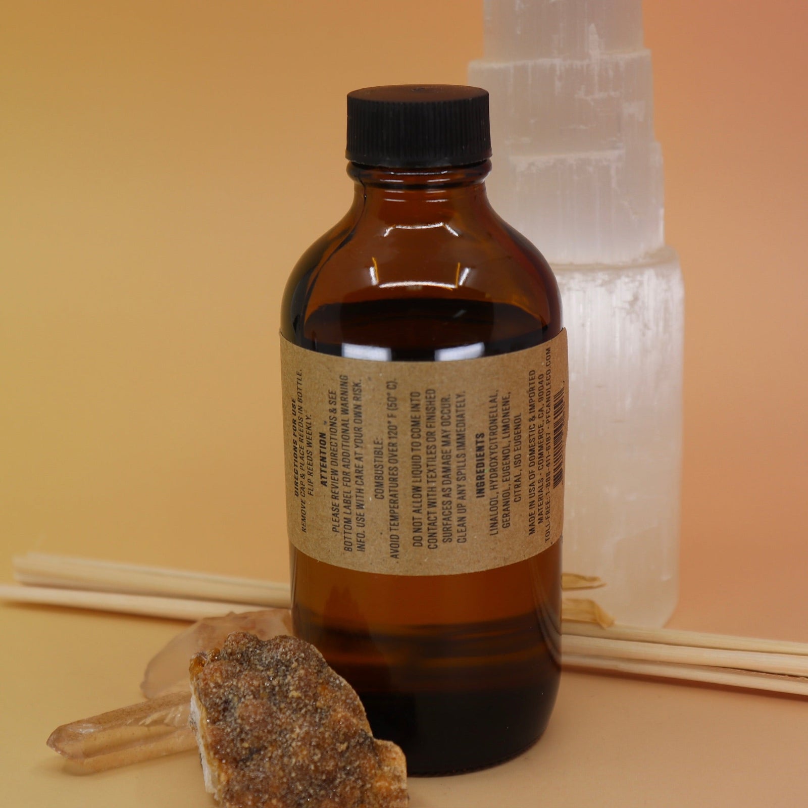 The back of the Amber & Moss - 3.5 oz Reed Diffuser - P.F. Candle Co. in front of a selenite tower all on a pink and yellow background.