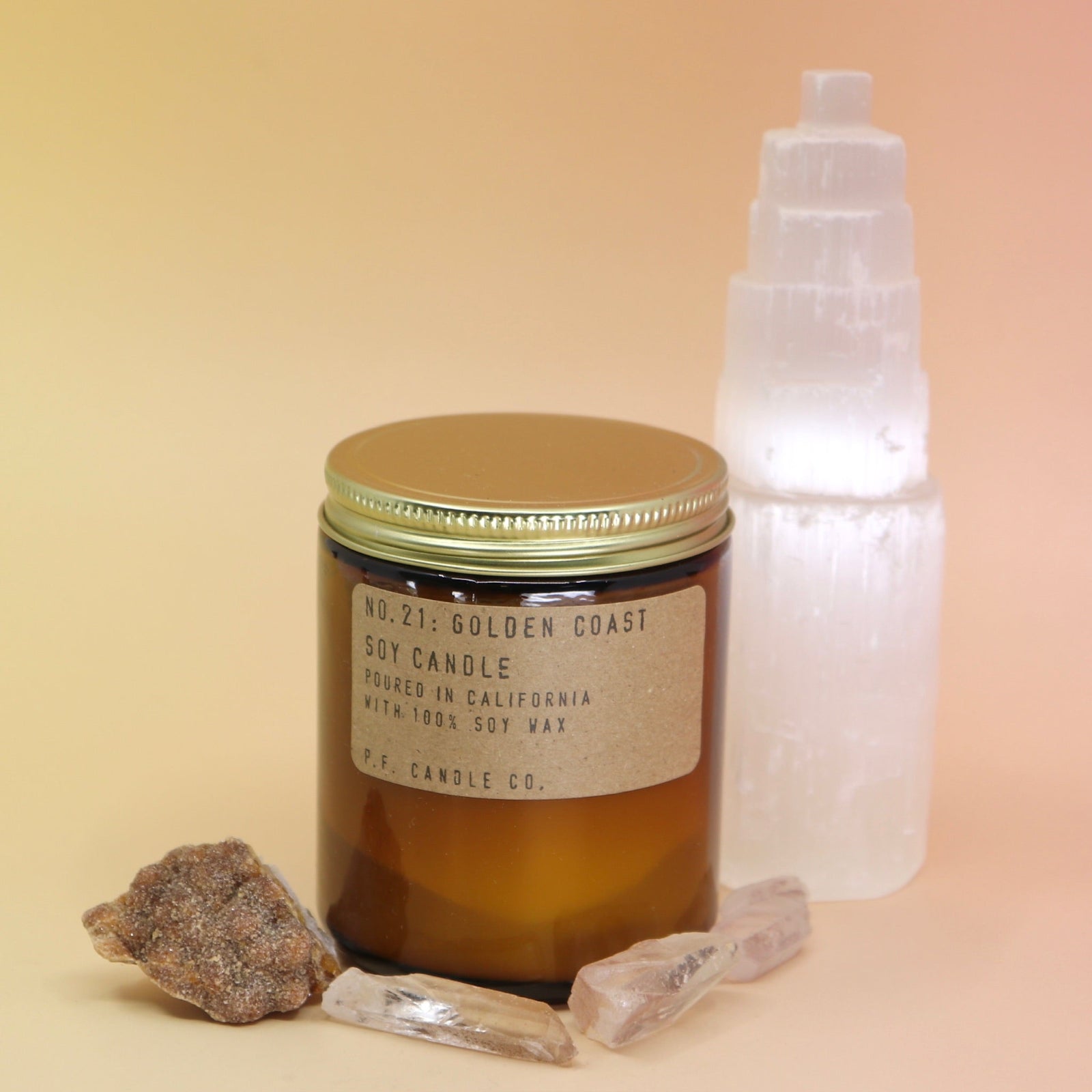 The Golden Coast - 7.2 oz Soy Wax Candle in front of a selenite tower surrounded by other crystals all on a pink and yellow background.