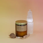 The Golden Coast - 7.2 oz Soy Wax Candle in front of a selenite tower surrounded by other crystals all on a pink and yellow background.