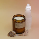 The Golden Coast - 7.2 oz Soy Wax Candle in front of a selenite tower surrounded by other crystals all on a pink and yellow background.
