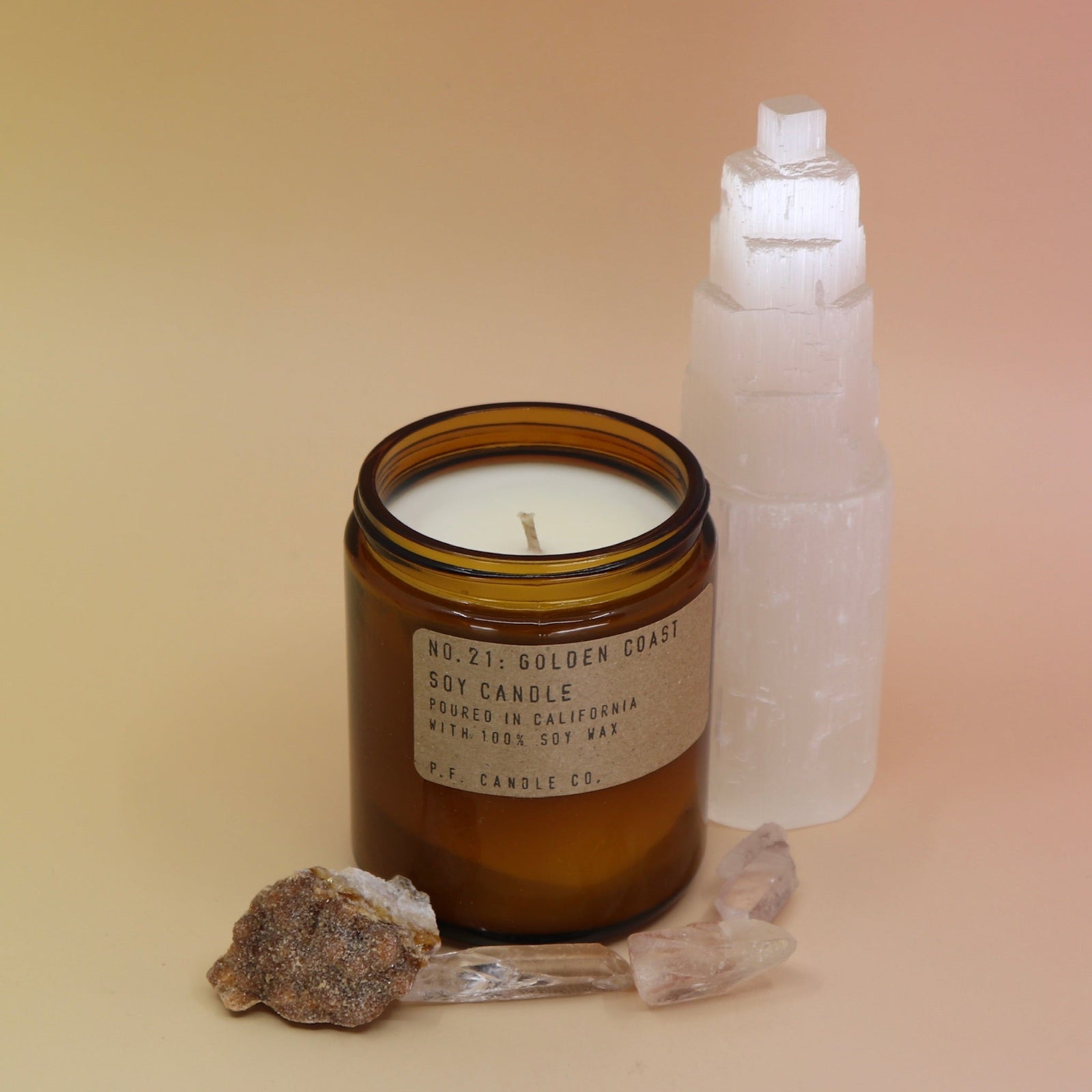 The Golden Coast - 7.2 oz Soy Wax Candle in front of a selenite tower surrounded by other crystals all on a pink and yellow background.