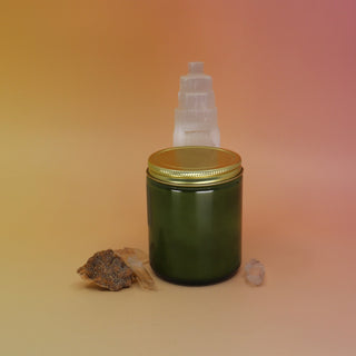 The P.F. Candle Co. Campfire 7.2 oz Standard Candle in front of a selenite tower on a pink and yellow background.
