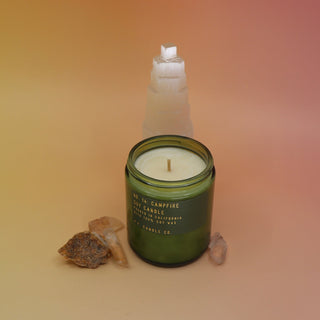 The P.F. Candle Co. Campfire 7.2 oz Standard Candle in front of a selenite tower on a pink and yellow background.
