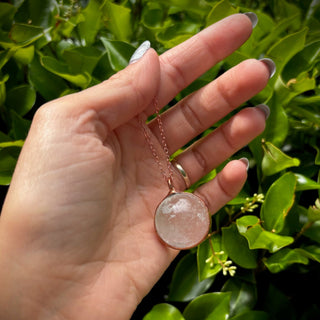 Someone holding the Crystal Ball Necklace in Copper - Clear Quartz in front of greenery.