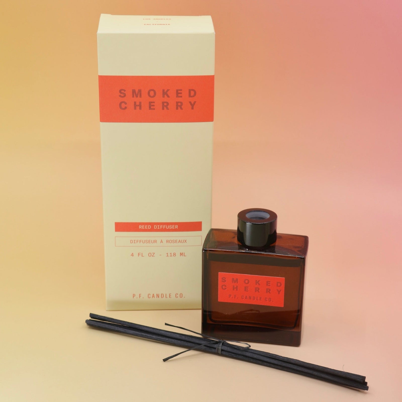 The Smoked Cherry – HI-FI Reed Diffuser next to its packaging with its reed sticks in front of it all on a pink and yellow background.