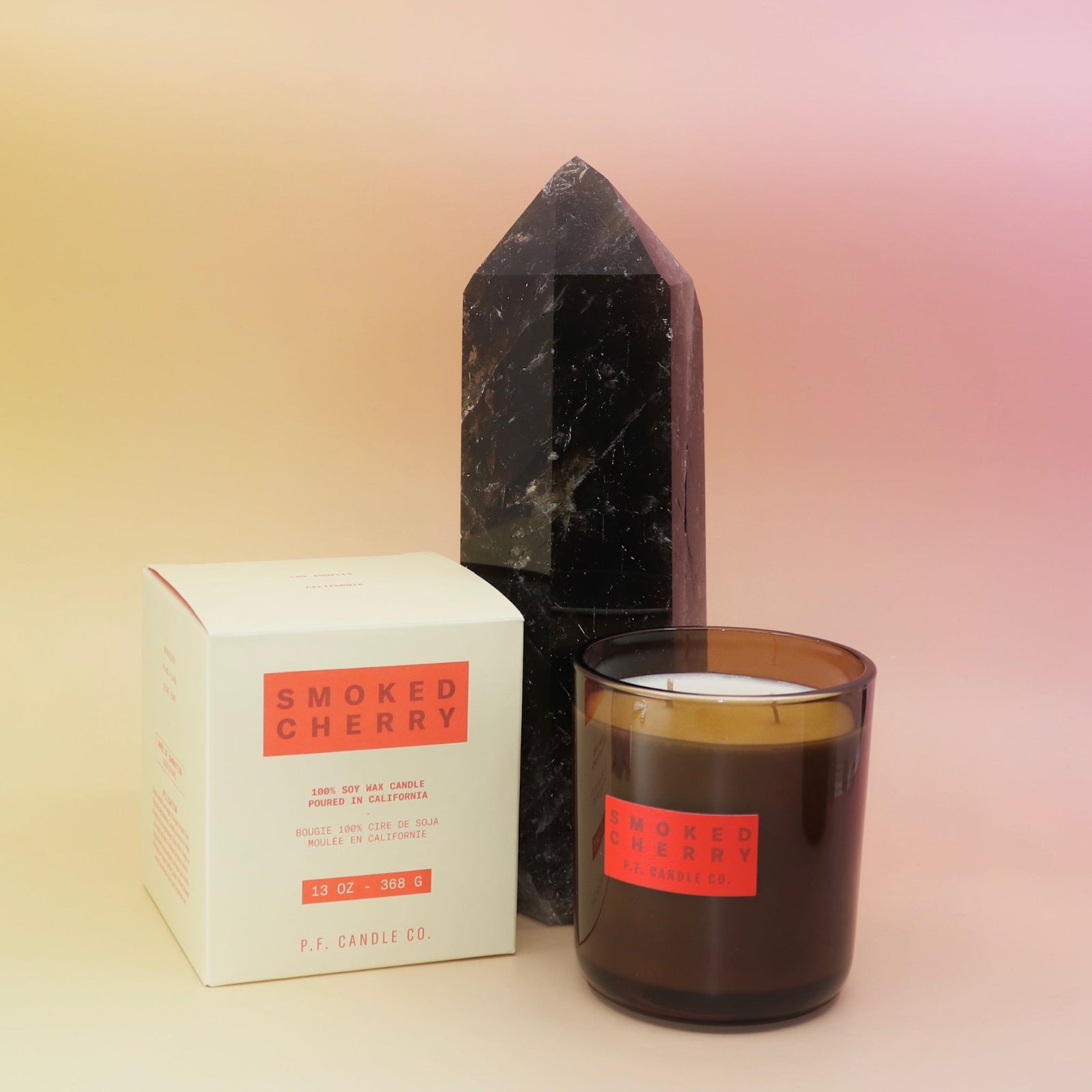 The Smoked Cherry - HI-FI Soy Wax Candle next to its packaging and in front of a crystal tower all on a pink and yellow background.