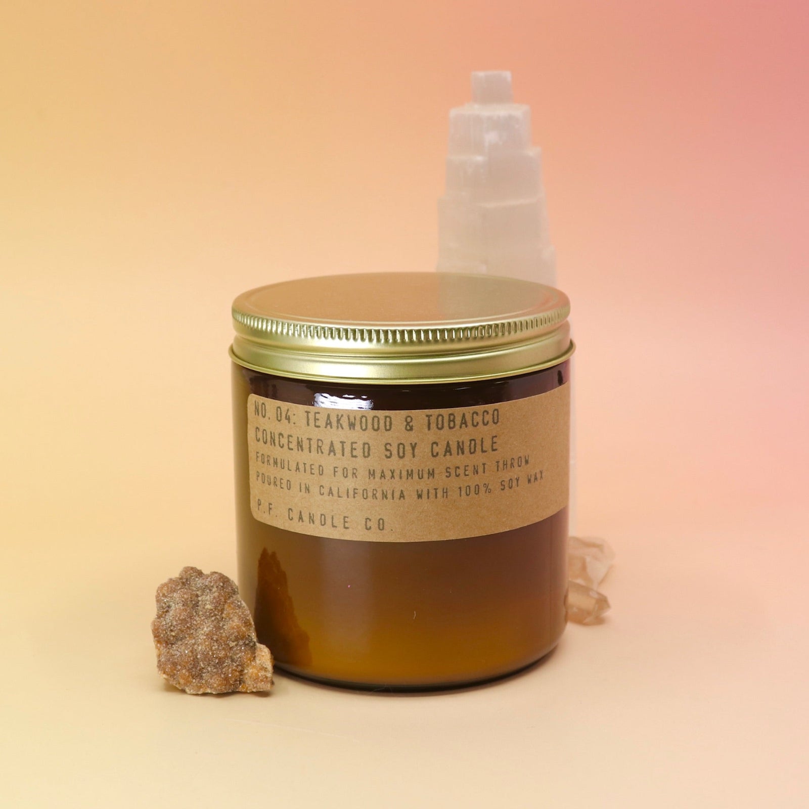 The P.F. Candle Co. Teakwood & Tobacco Large Candle in front of a selenite tower on a pink and yellow background.