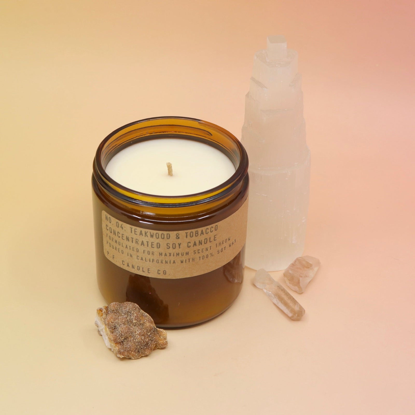 The P.F. Candle Co. Teakwood & Tobacco Large Candle in front of a selenite tower on a pink and yellow background.