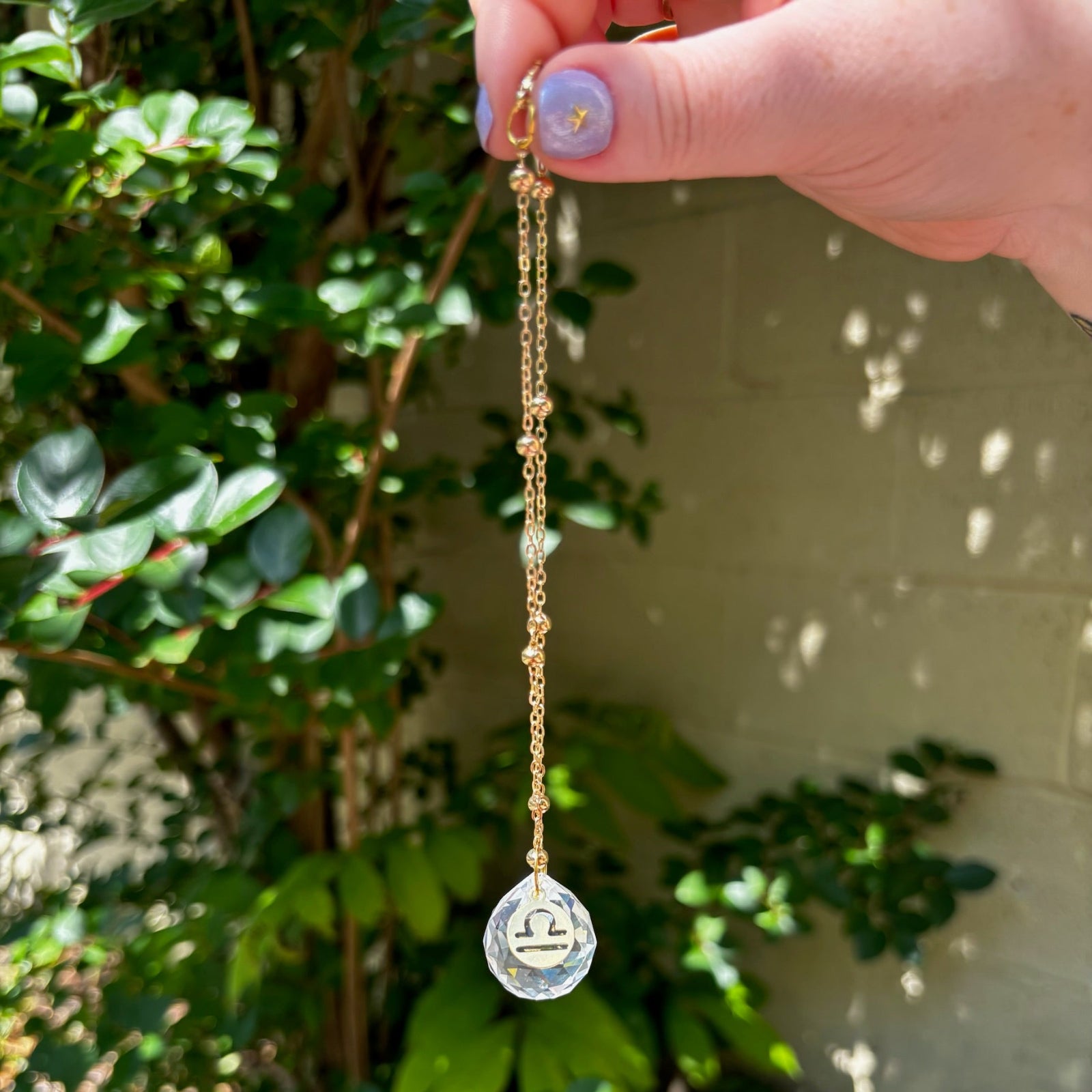 The Libra Zodiac Car Charm Suncatcher being held in front of a cream-colored brick wall with greenery.