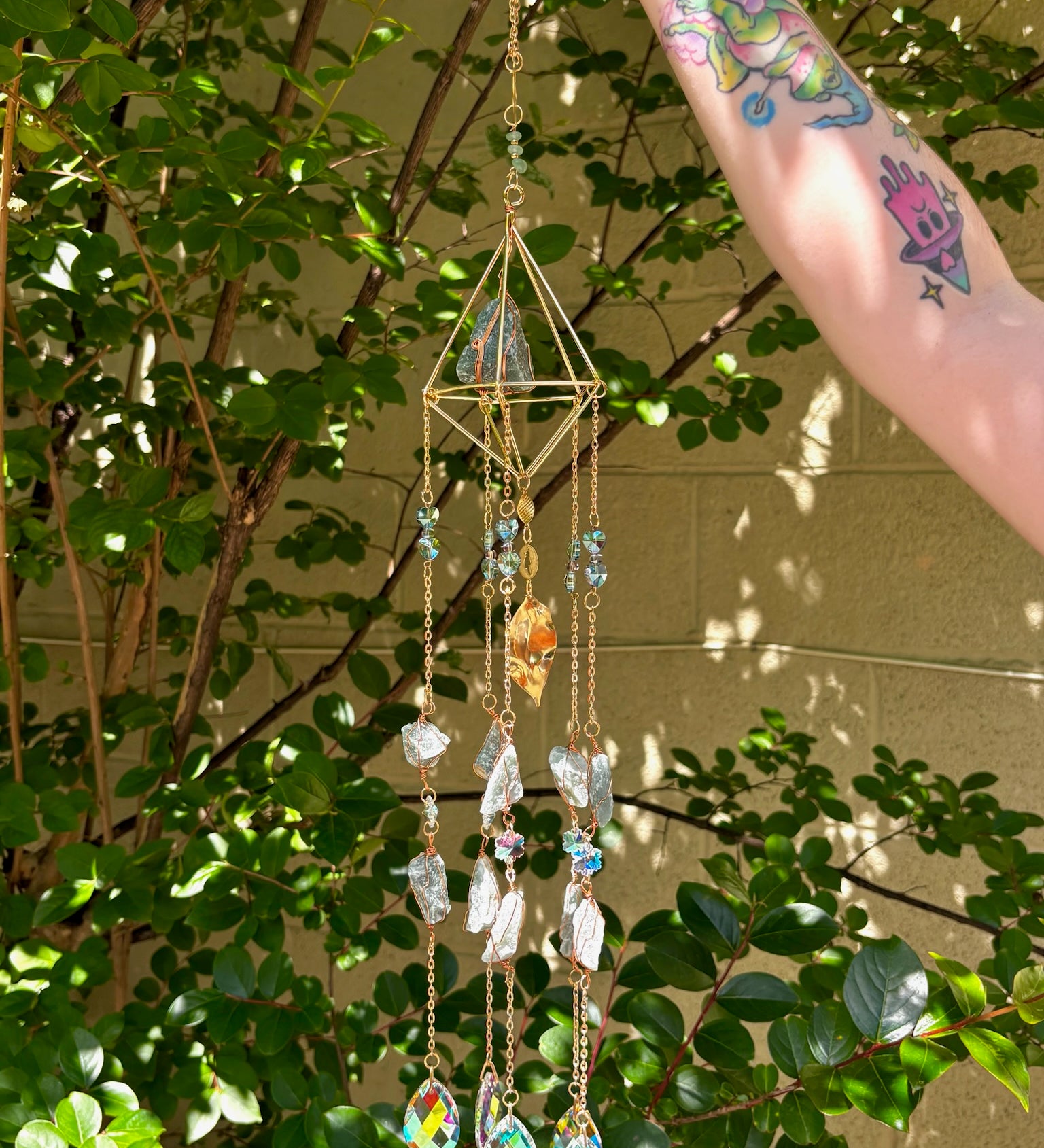 Someone holding the Amazonite Lotus Crystal Suncatcher in front of a cream-colored brick wall with greenery.
