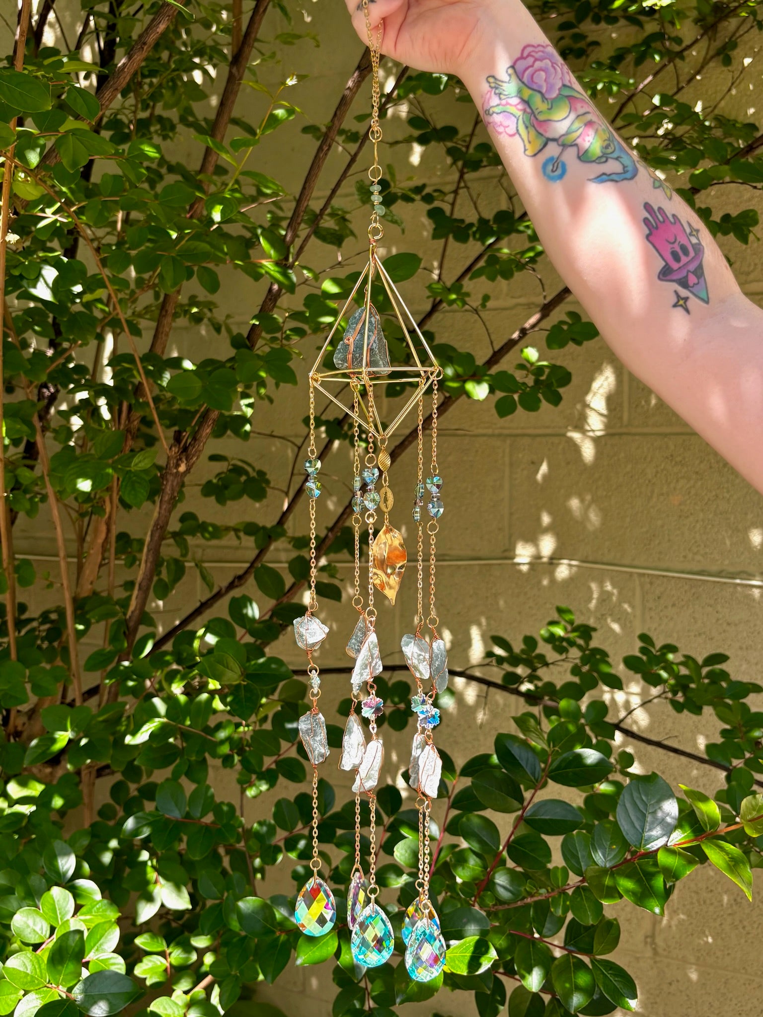 Someone holding the Amazonite Lotus Crystal Suncatcher in front of a cream-colored brick wall with greenery.