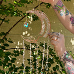Someone holding the rose quartz Moon Crystal Gemstone Suncatcher in front of a cream-colored brick wall with greenery.