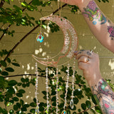 Someone holding the rose quartz Moon Crystal Gemstone Suncatcher in front of a cream-colored brick wall with greenery.