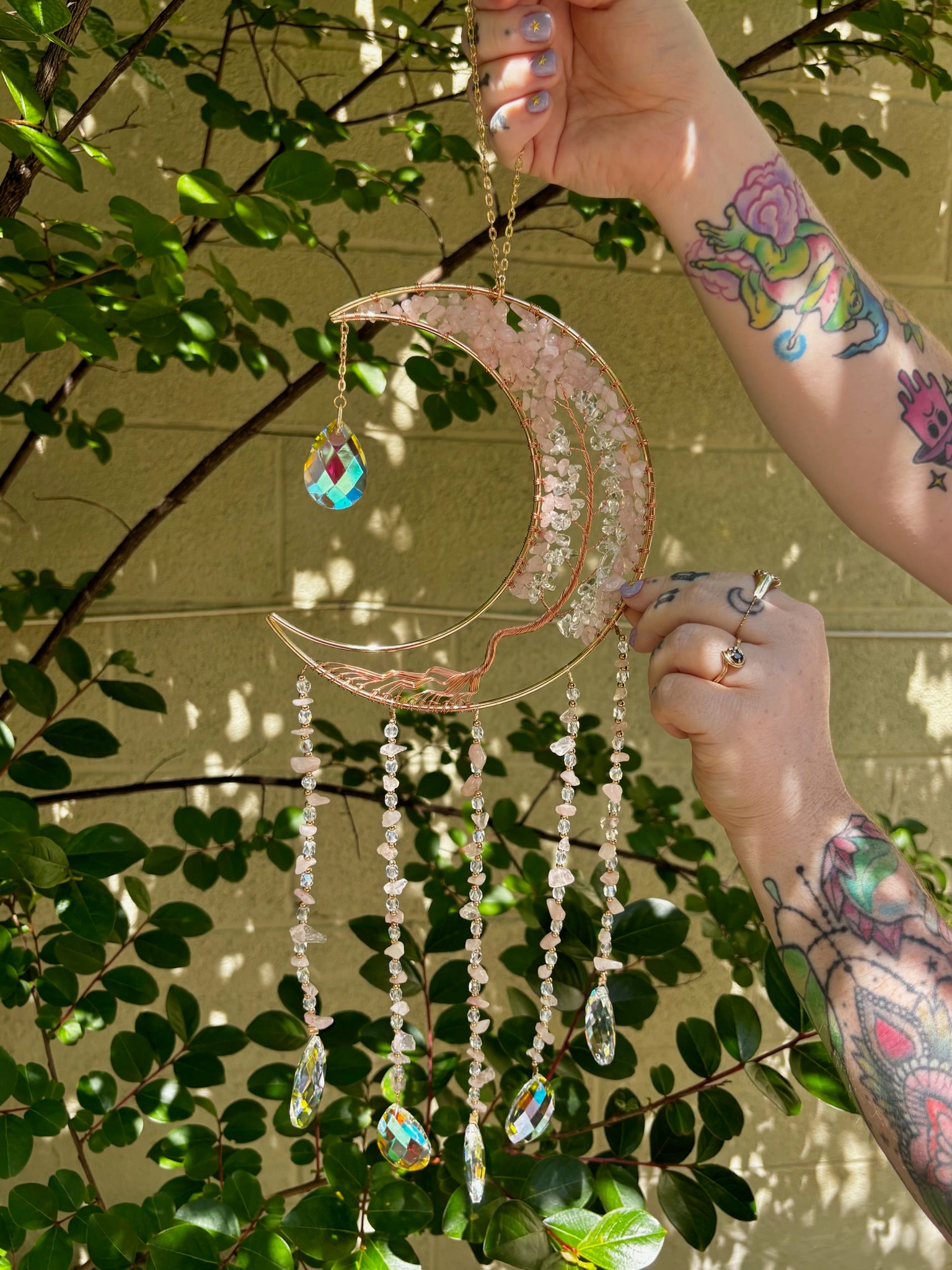 Someone holding the rose quartz Moon Crystal Gemstone Suncatcher in front of a cream-colored brick wall with greenery.